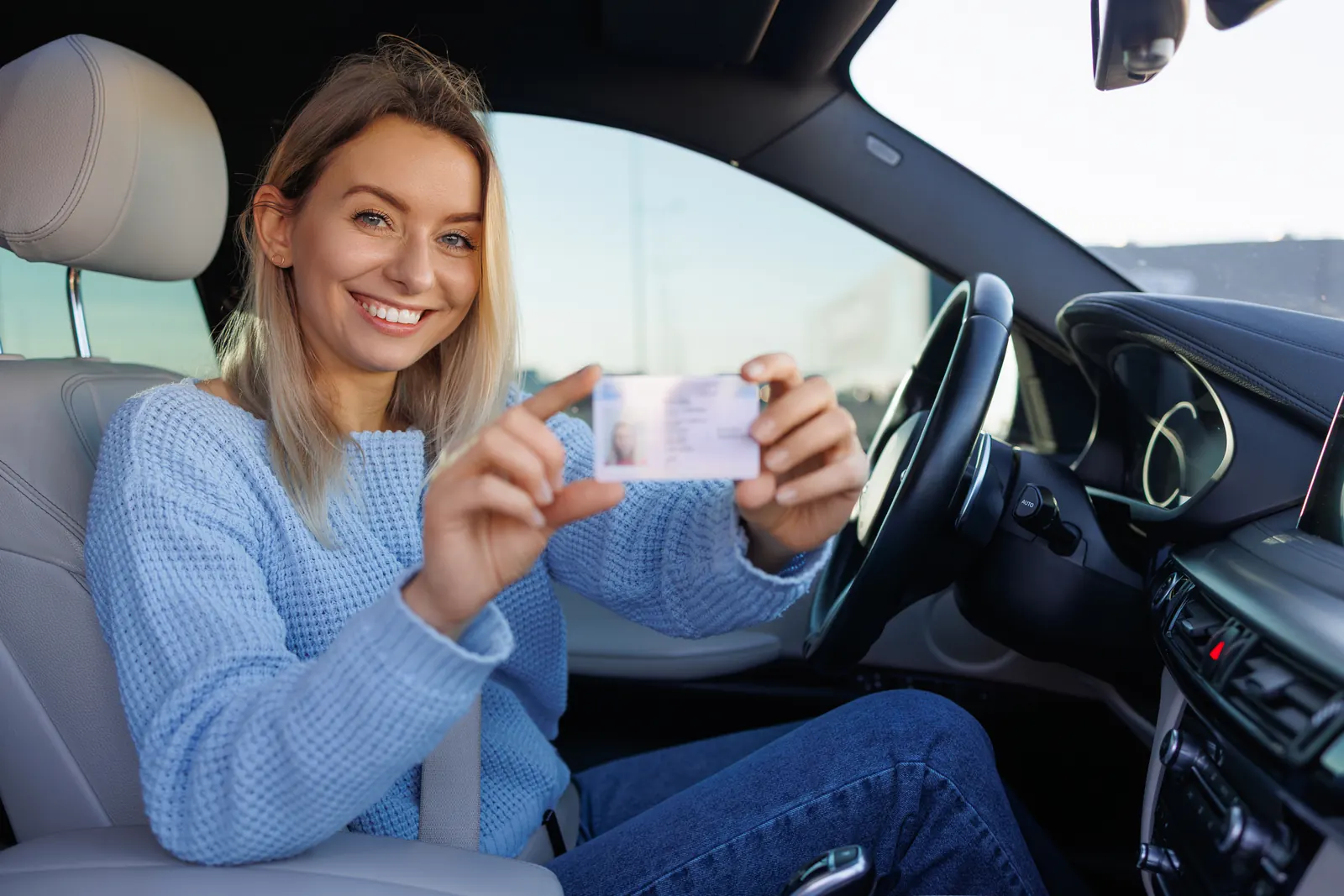 Junge Frau sitzt am Steuer und zeigt lächelnd ihren neuen Führerschein